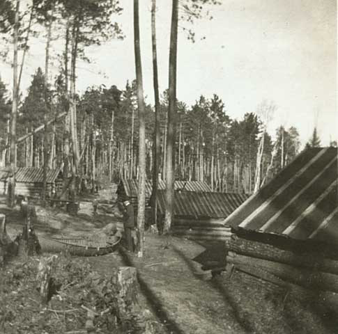 Landscape dominated by a canoe, standing man, trees, and side of a building in the right foreground, more buildings in the middleground, and trees in the background.
